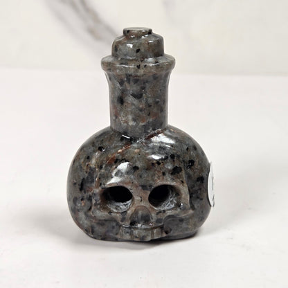 A small skull-shaped bottle or pendant made of polished dark stone with black, gray, and brown speckling, shown on a plain light background.