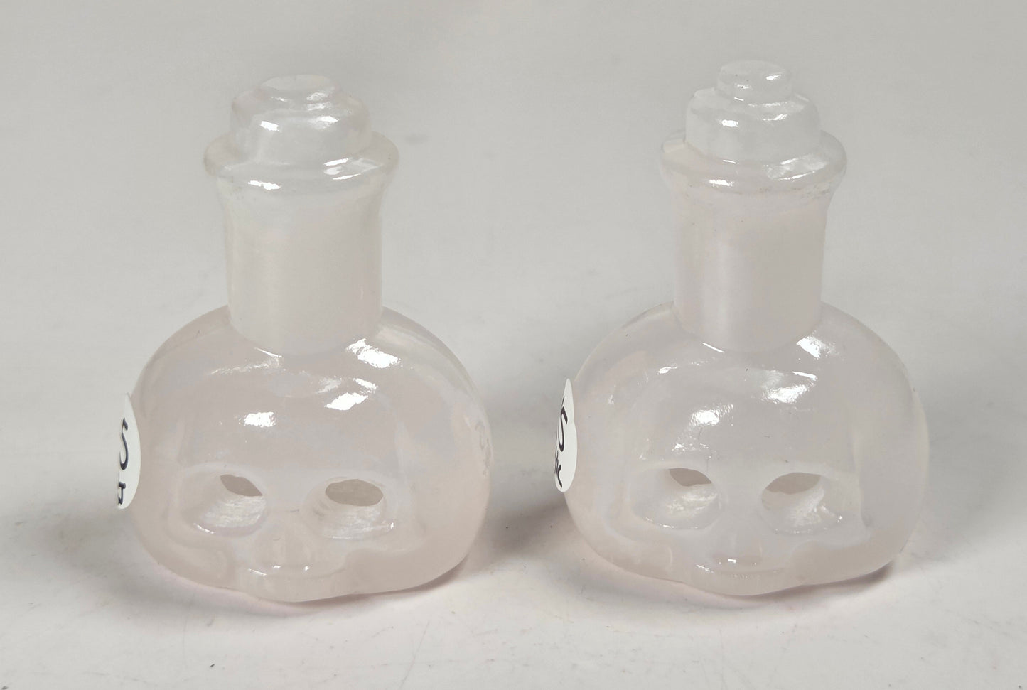 A pair of small translucent glass bottles shaped like skulls, with narrow necks and stoppers, photographed on a plain light background.