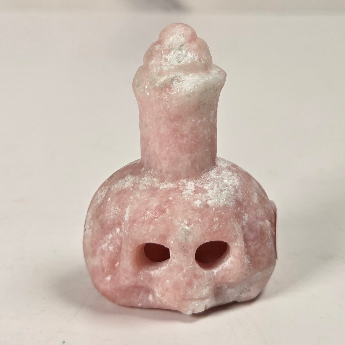 A small pink stone object with a rounded body, two dark eye openings, and a narrow neck topped with a textured, frosted cap, photographed on a plain light background.