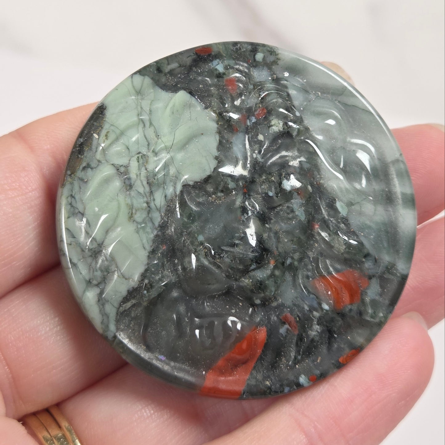 A hand holding a round polished stone pendant featuring a carved lion design, with natural marbled patterns in gray, black, pale green, and orange-red.