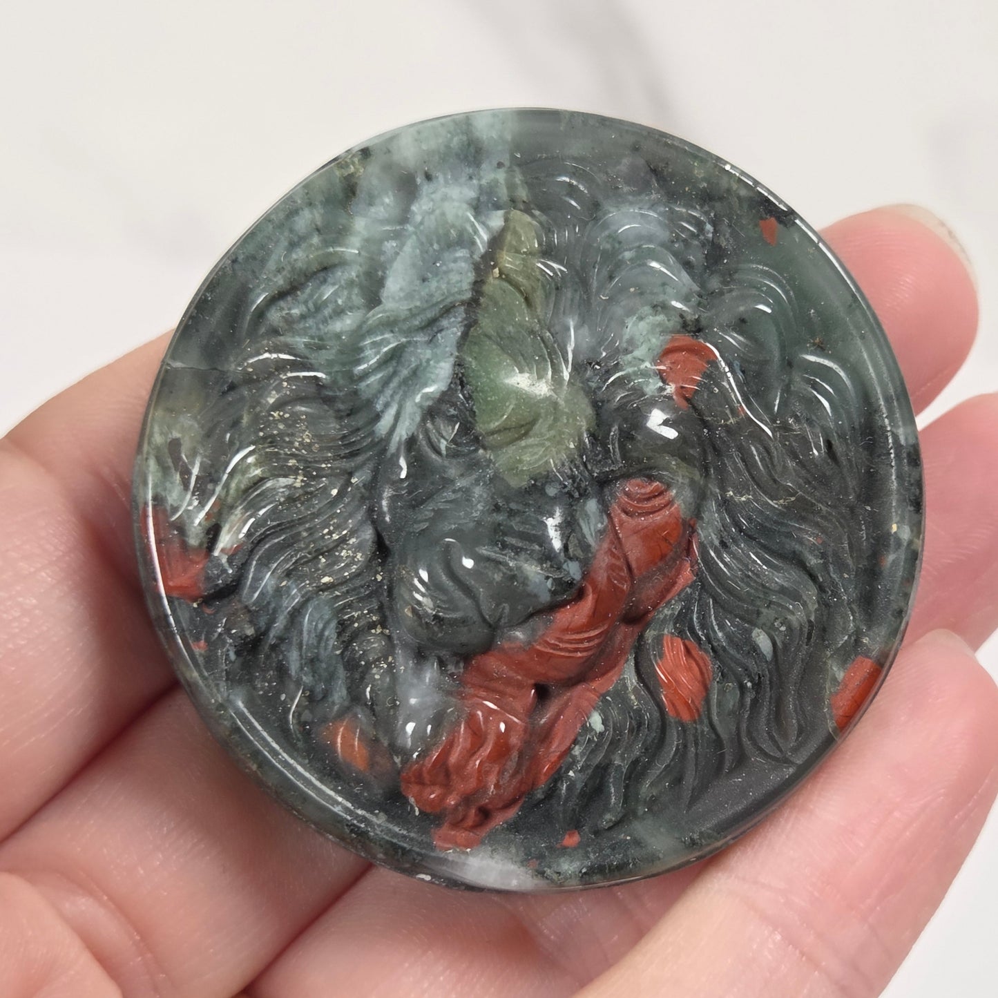 A hand holds a round carved stone pendant featuring a lion head with gray, green, and rust-red marbled details.