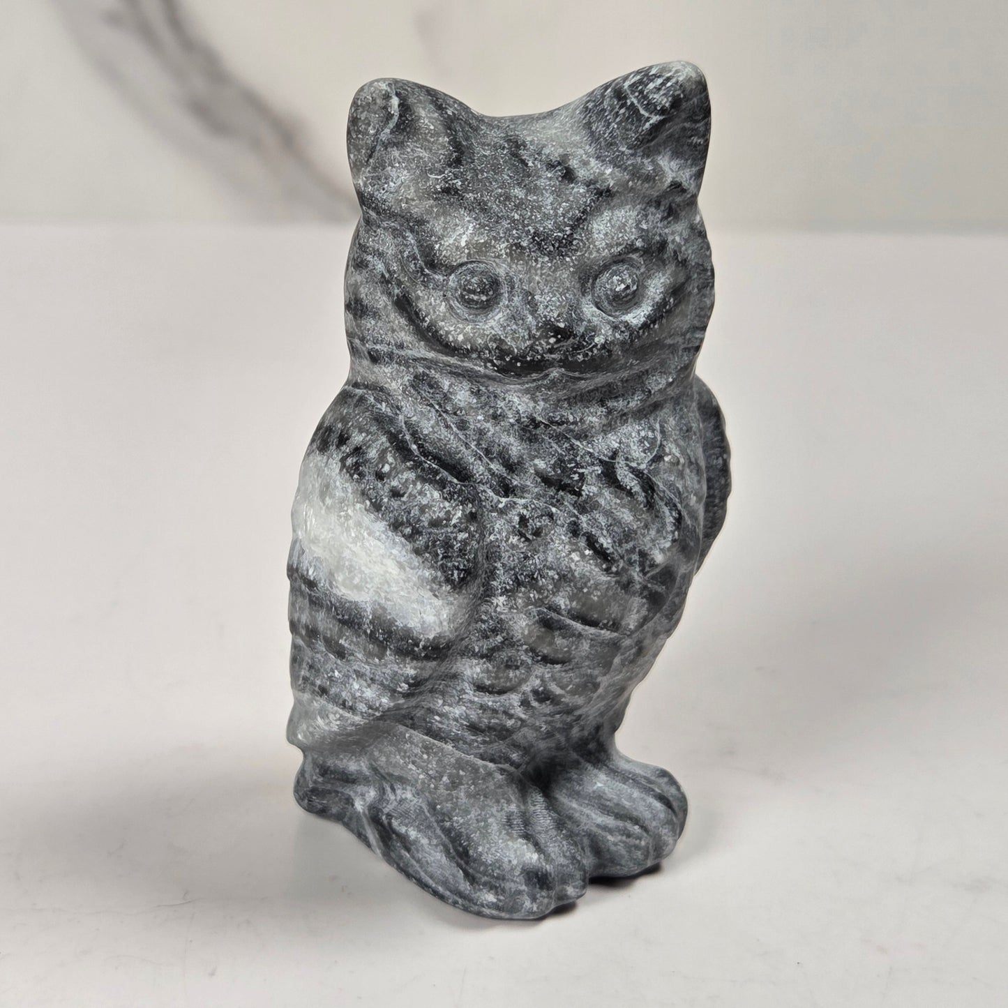 A small carved stone owl figurine in dark gray and white marbled stone, standing upright on a light gray surface.