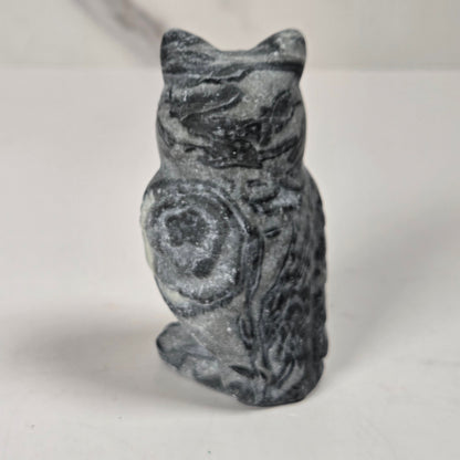 A small carved stone owl figurine with a mottled dark gray and white pattern, standing upright on a plain light surface.