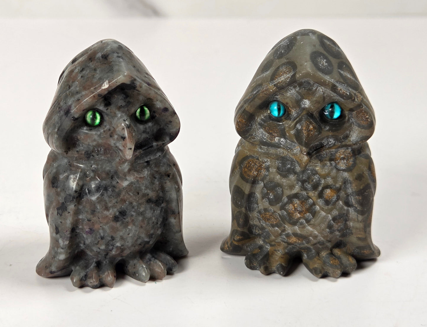 Two small owl figurines made from polished stone, one dark gray with green eyes and the other olive green with blue eyes, displayed side by side on a white surface.