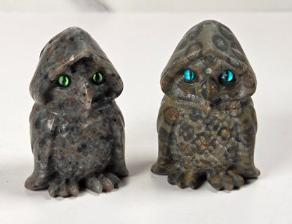 Two small owl figurines made from polished stone, one dark gray with green eyes and the other olive green with blue eyes, displayed side by side on a white surface.