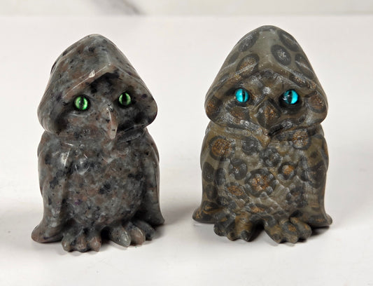 Two small owl figurines made from polished stone, one dark gray with green eyes and the other olive green with blue eyes, displayed side by side on a white surface.