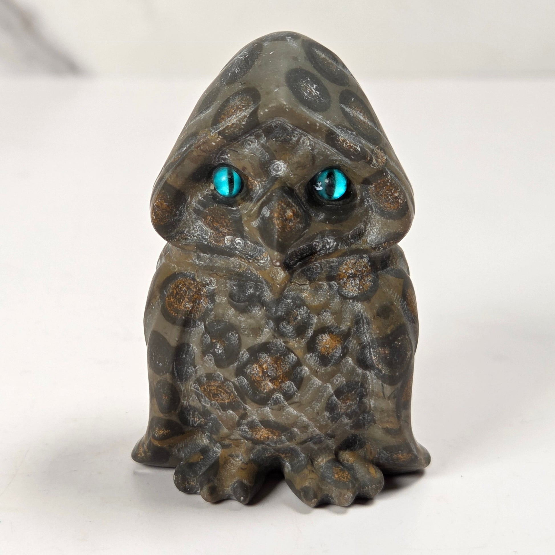 A small owl figurine with a textured, mottled gray and brown stone-like appearance and glowing turquoise-blue eyes, standing on a white surface.