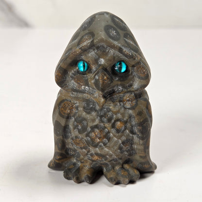 A small owl figurine with a textured, mottled gray and brown stone-like appearance and glowing turquoise-blue eyes, standing on a white surface.