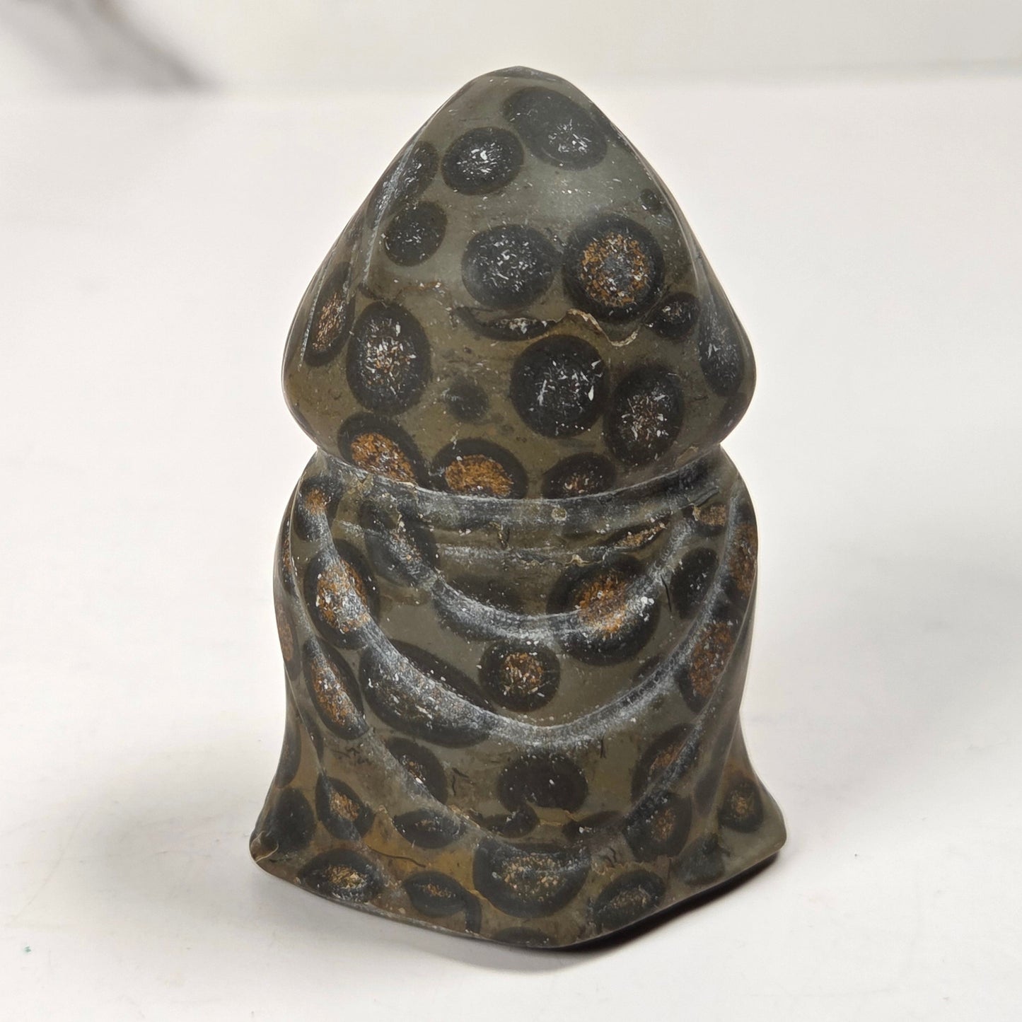 A small sculpted figurine of a hooded robed figure, likely representing a saint or monk, with a textured surface covered in black circular spots with warm brown centers, set against a plain white background.