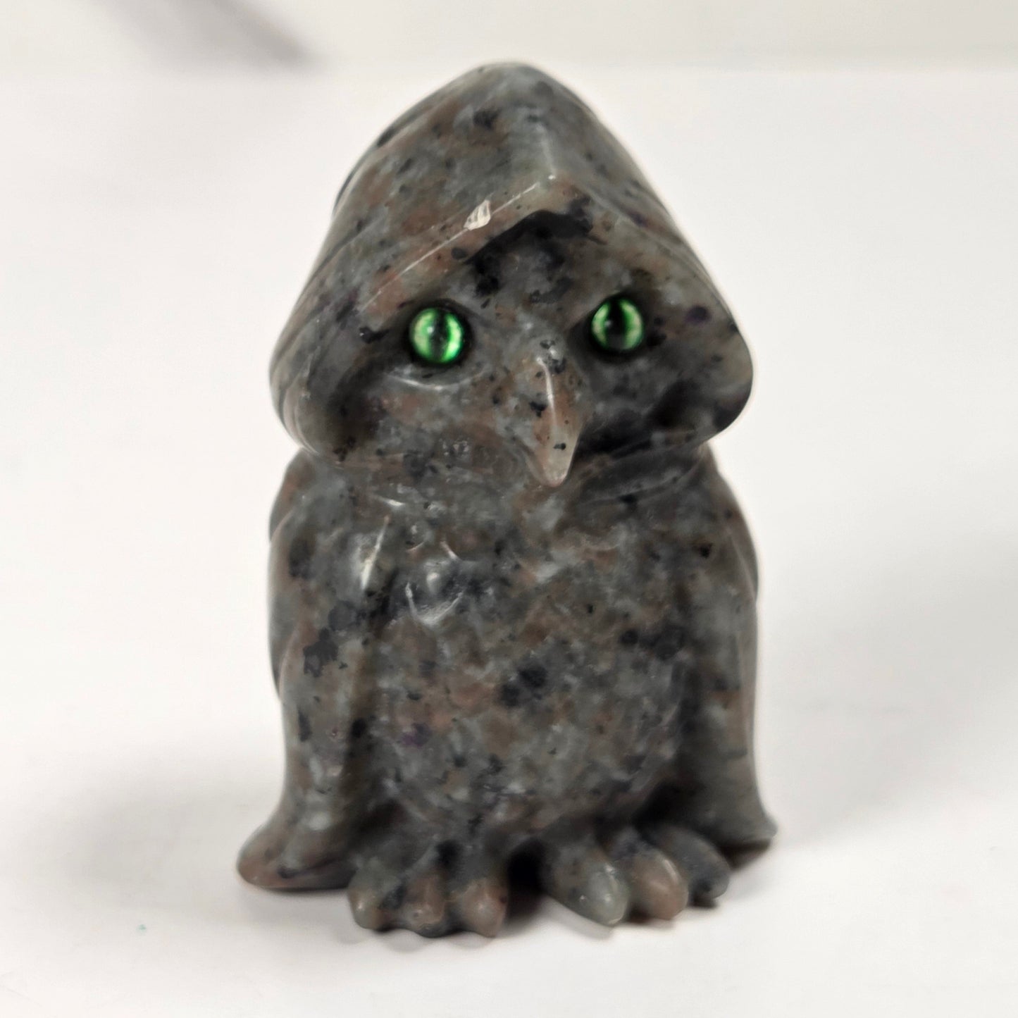 A small carved stone owl figurine with a hooded head and bright green eyes, standing on a plain light background.