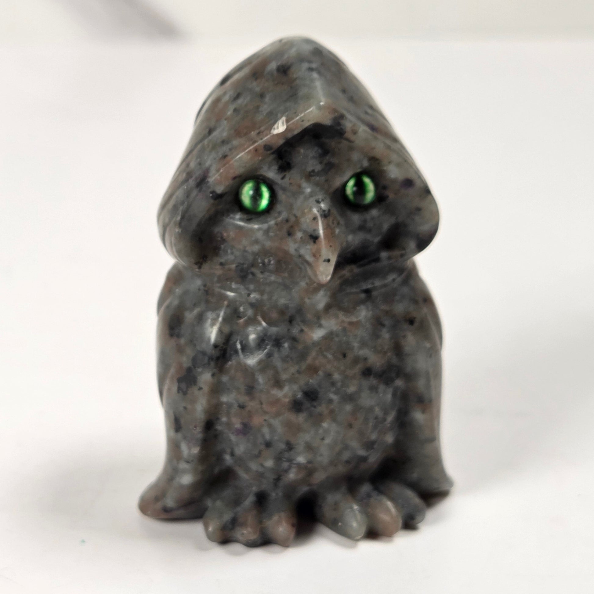 A small carved stone owl figurine with a hooded head and bright green eyes, standing on a plain light background.