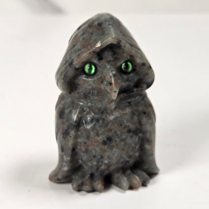 A small carved stone owl figurine with a hooded head and bright green eyes, standing on a plain light background.