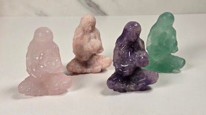Four small carved crystal figurines of seated meditating monks, made from rose quartz, pink tourmaline, amethyst, and green tourmaline, arranged on a white surface with a marble-patterned background.