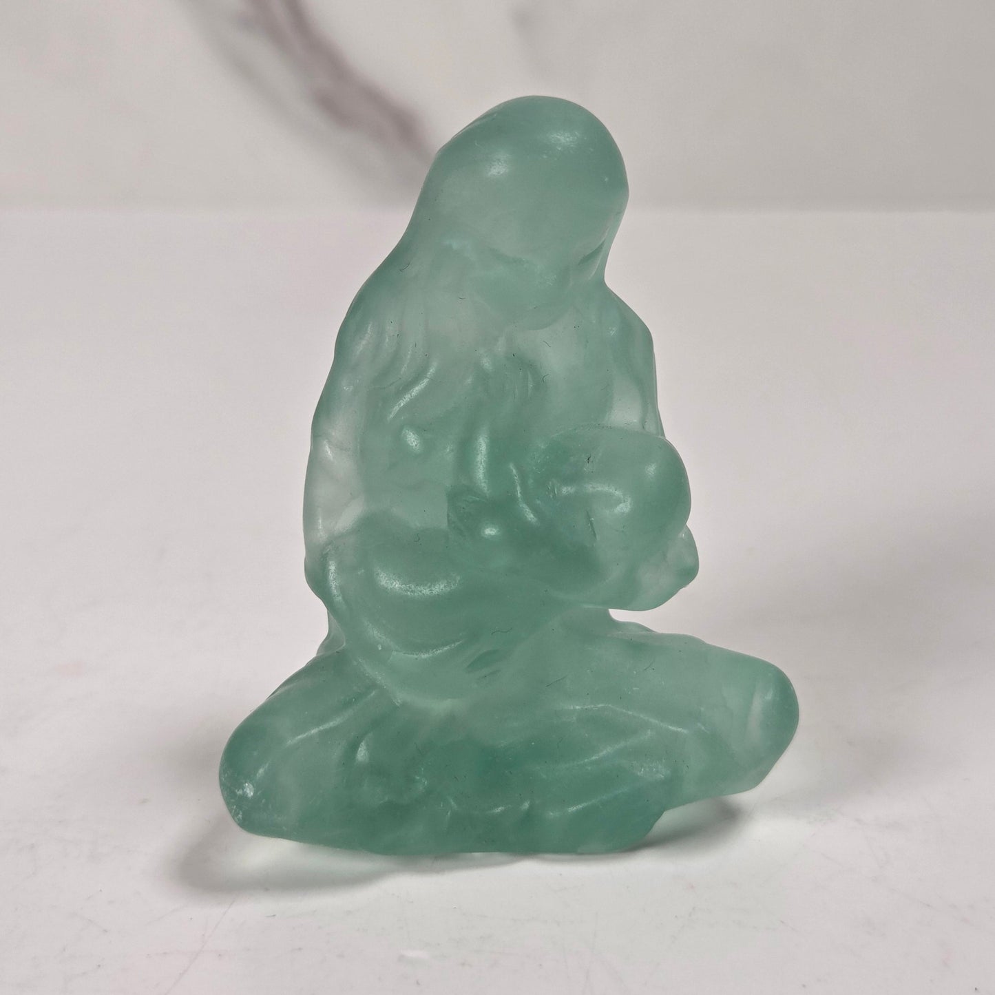 A small translucent green jade-like figurine of a seated Buddha, photographed on a light gray surface with a marble-patterned background.
