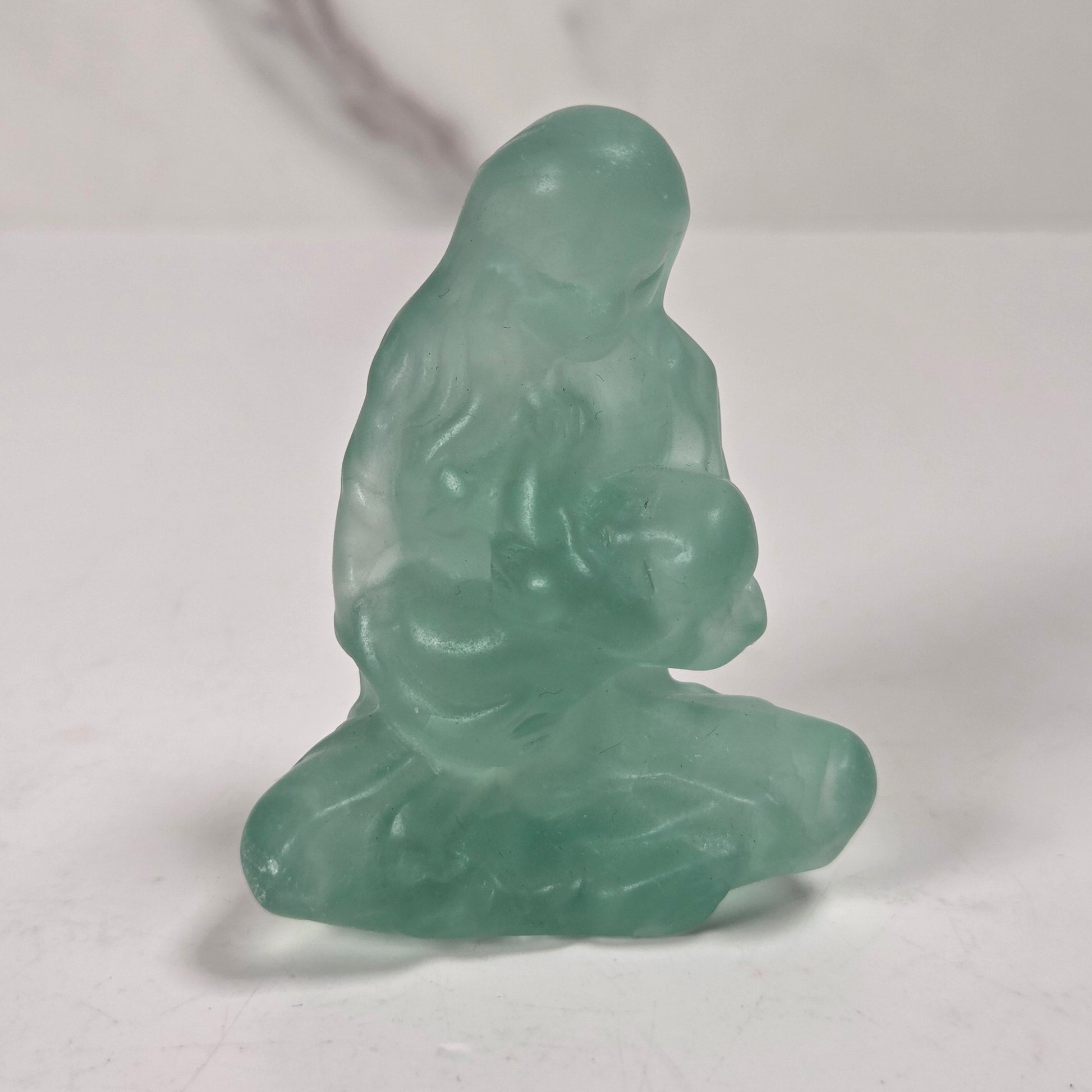 A small translucent green jade-like figurine of a seated Buddha, photographed on a light gray surface with a marble-patterned background.