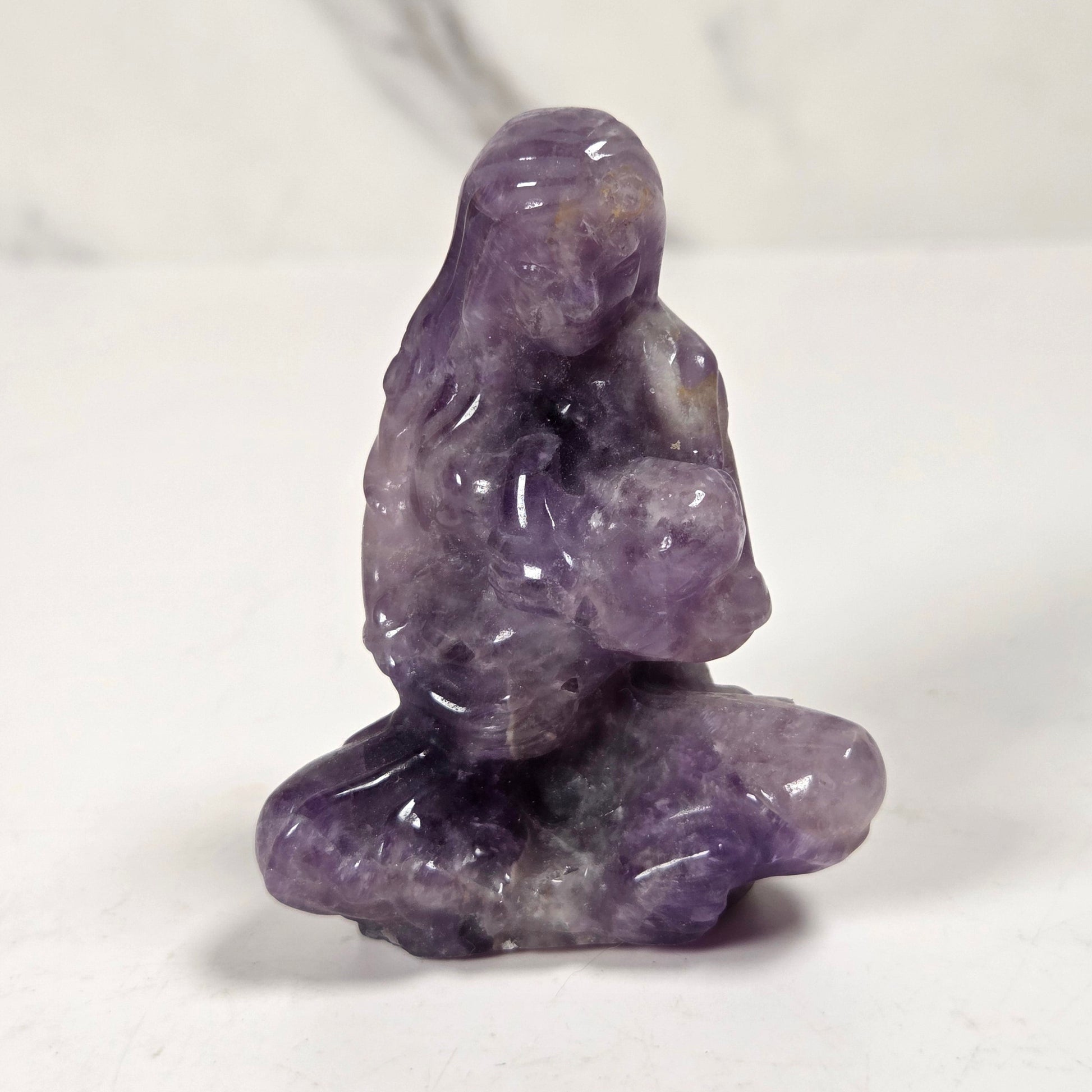 A small carved purple crystal figurine of a seated meditating figure, photographed on a white surface with a light gray marble-patterned background.