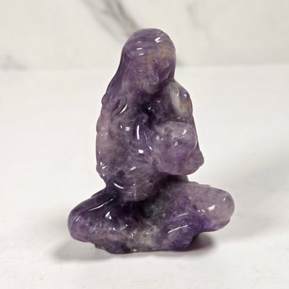 A small carved purple crystal figurine of a seated meditating figure, photographed on a white surface with a light gray marble-patterned background.