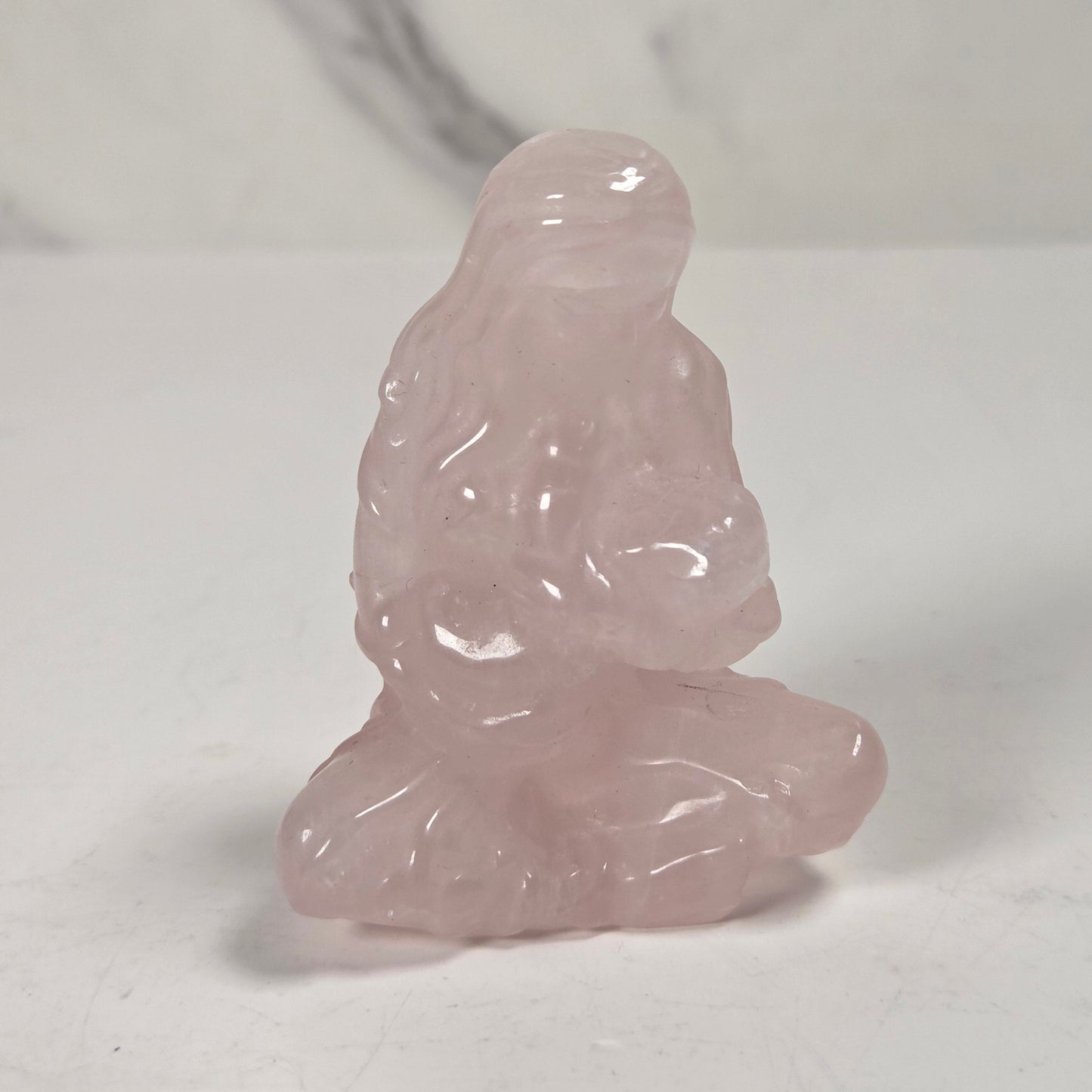 A small pink translucent figurine carved in a seated Buddha-like pose, photographed on a light neutral surface with a soft shadow.