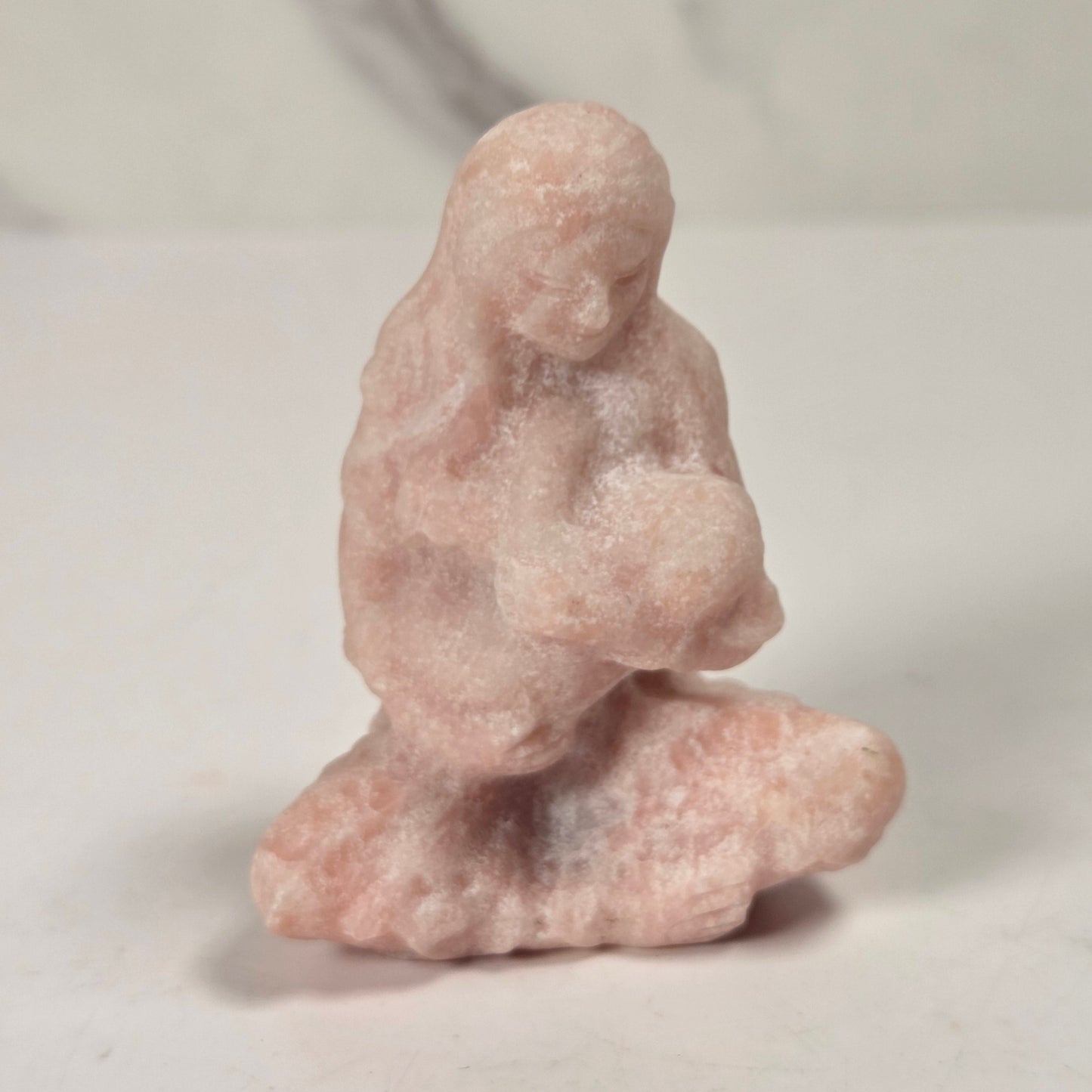 A small pink stone figurine of a kneeling, crouched human figure, photographed on a light surface with a soft marble-like background.