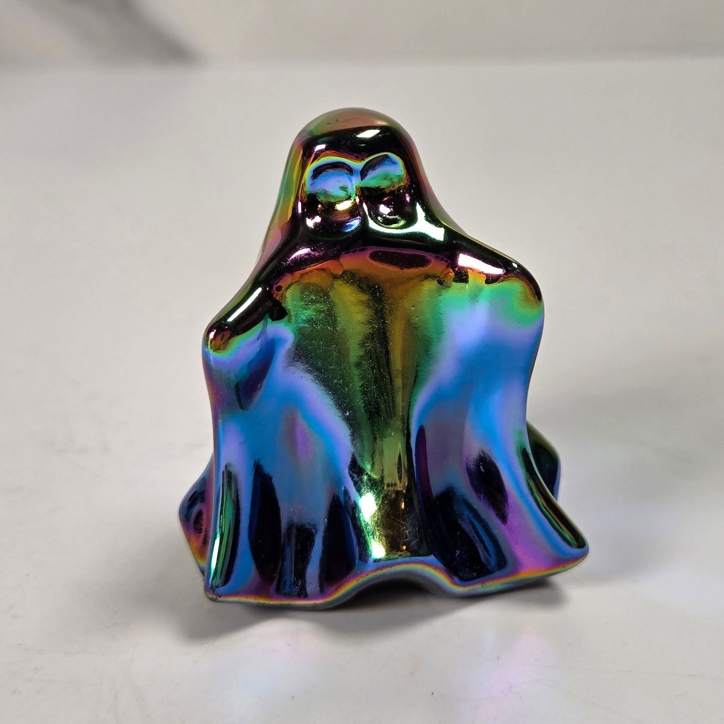 A small ghost-shaped figurine with a glossy, iridescent rainbow finish, standing on a light gray surface.