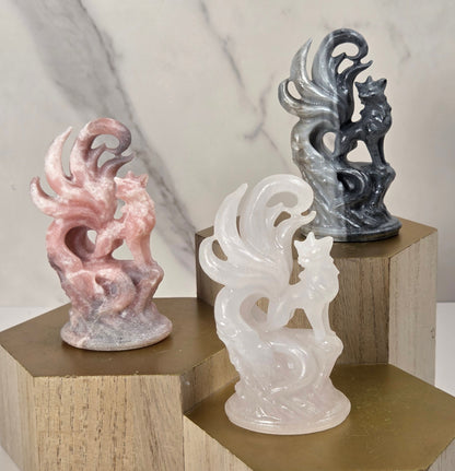 Three small carved figurines displayed on hexagonal wooden pedestals against a light marble-look background. The left figurine is pink with gray shading, the center figurine is translucent milky white, and the right figurine is dark gray with black shading.