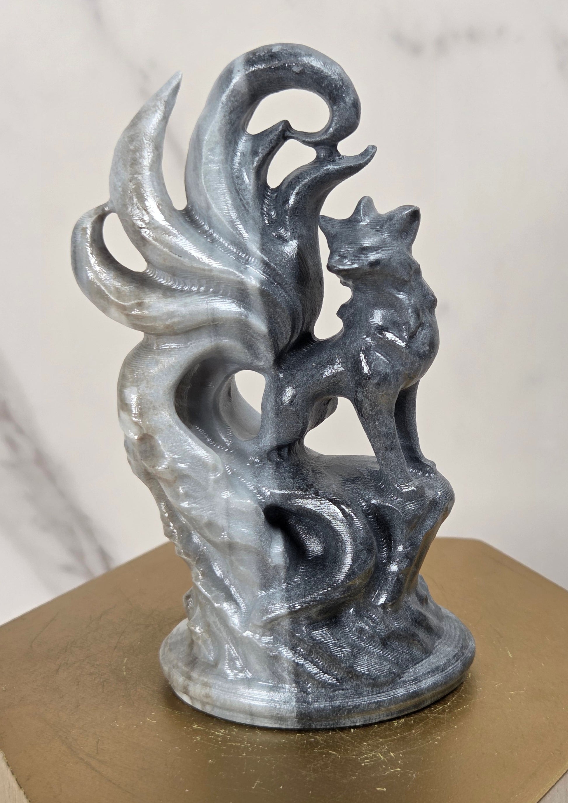 A small gray and black marbled figurine of a fox standing on a swirling flame-like base, displayed on a gold hexagonal pedestal against a light marble-patterned background.
