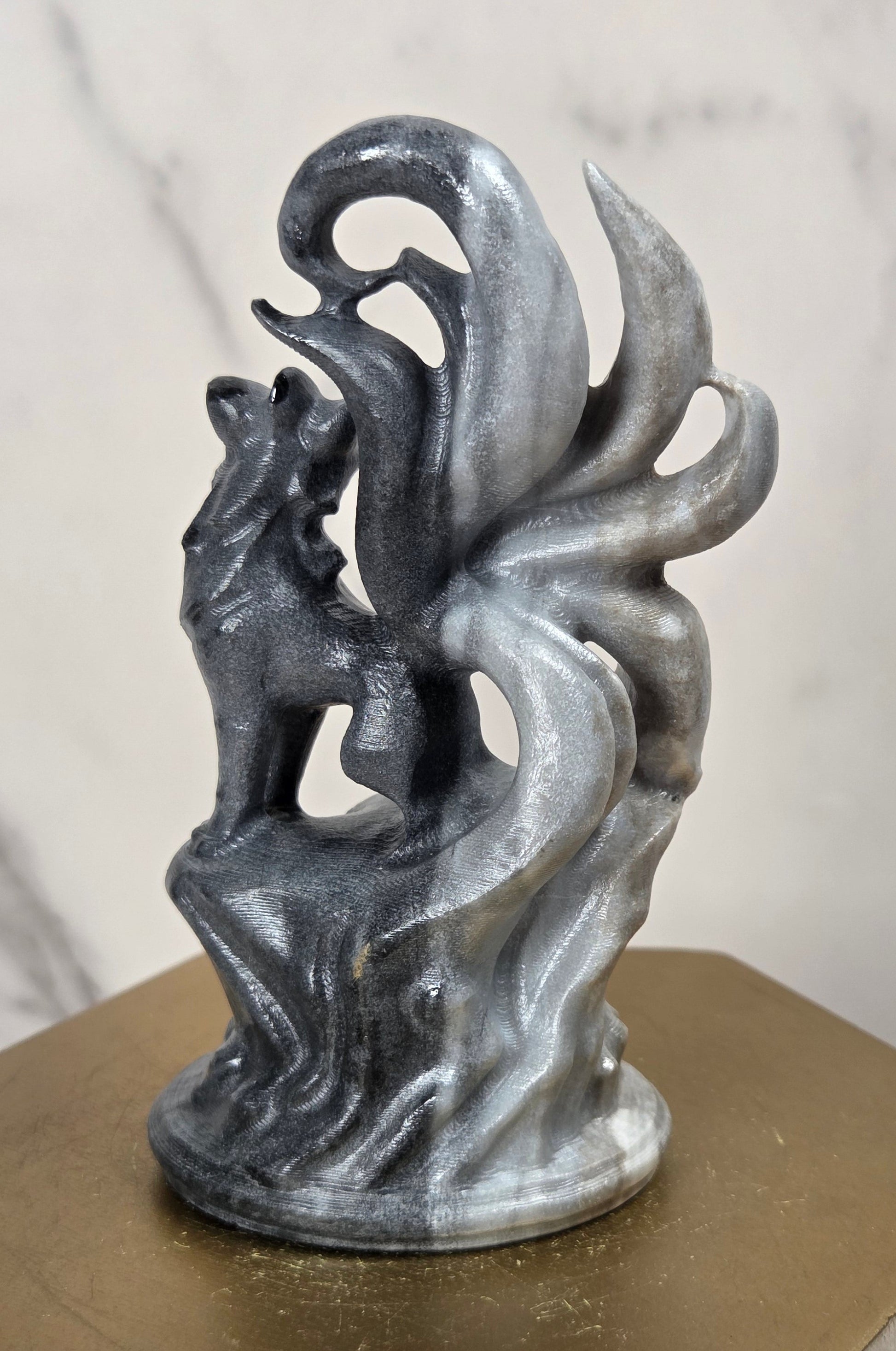 A metallic silver-gray sculpture of a fox with flowing tail-like forms, displayed on a hexagonal gold-toned pedestal against a light marble-patterned background.