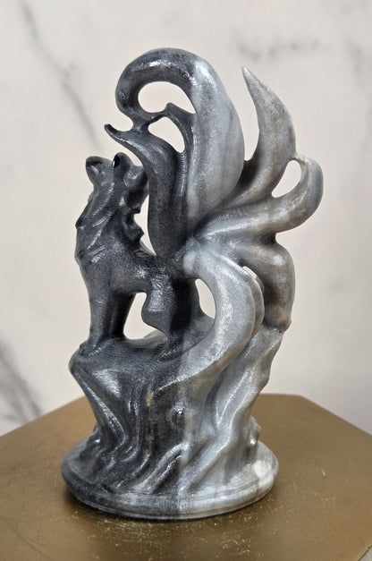 A metallic silver-gray sculpture of a fox with flowing tail-like forms, displayed on a hexagonal gold-toned pedestal against a light marble-patterned background.