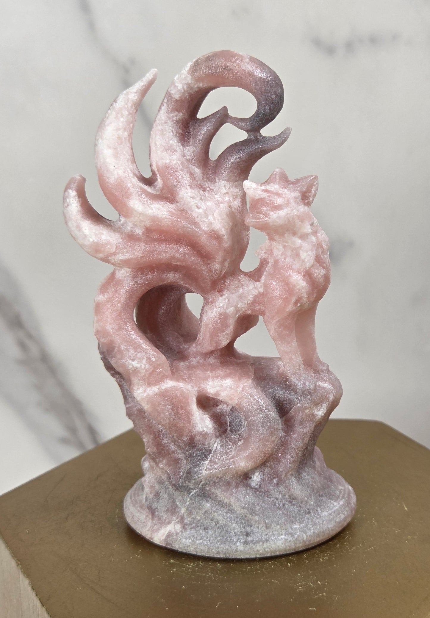 A pink stone figurine of a fox with flowing tail-like curls, displayed on a hexagonal gold pedestal against a light marble-patterned background.
