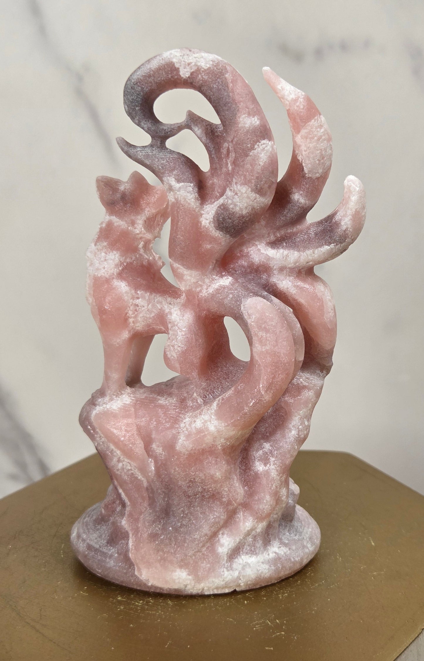 A small abstract pink sculpture with a textured, stone-like finish, displayed on a hexagonal gold pedestal against a light gray marble-patterned background.