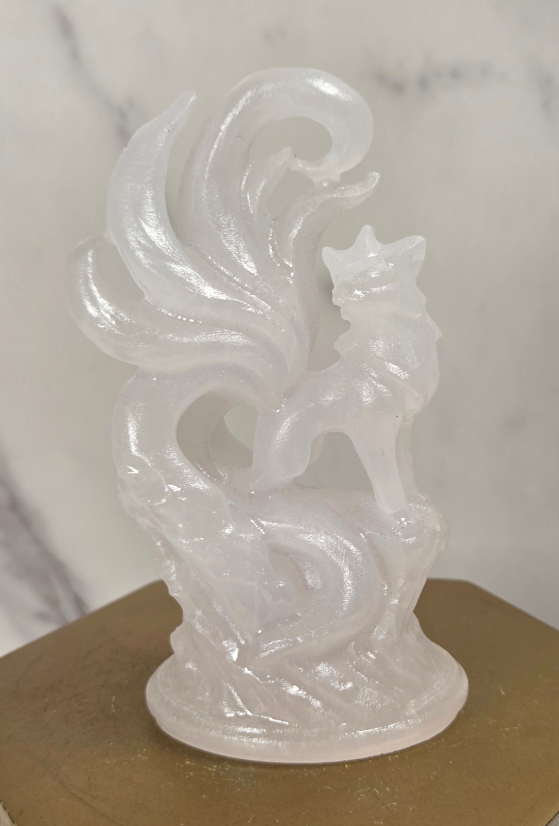 A translucent white carved figurine of a fox or wolf standing on a base with flowing tail-like forms, displayed on a gold-toned hexagonal pedestal against a light marble-patterned background.
