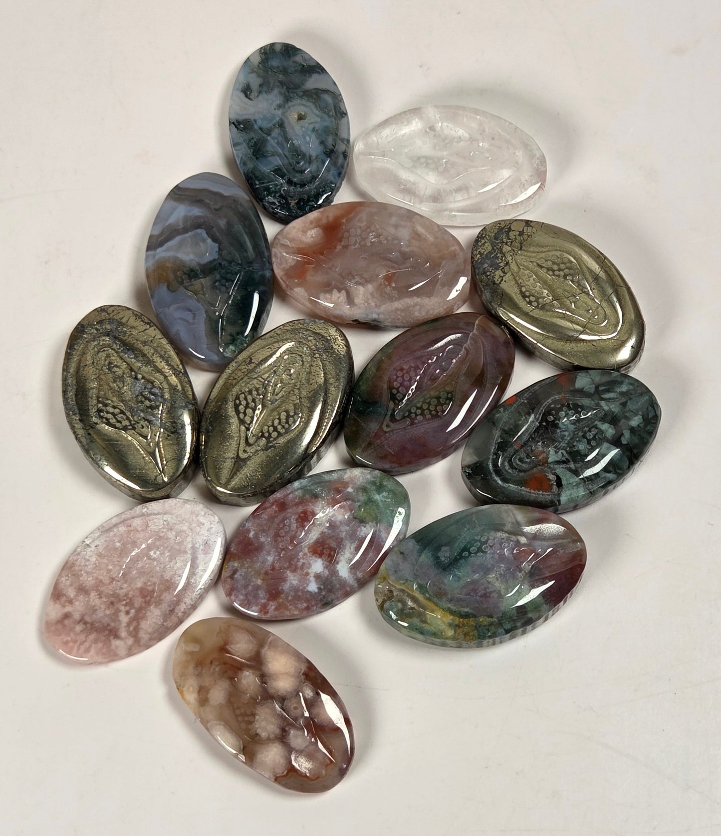 A group of polished oval gemstone cabochons in various colors and patterns, arranged on a plain off-white surface.