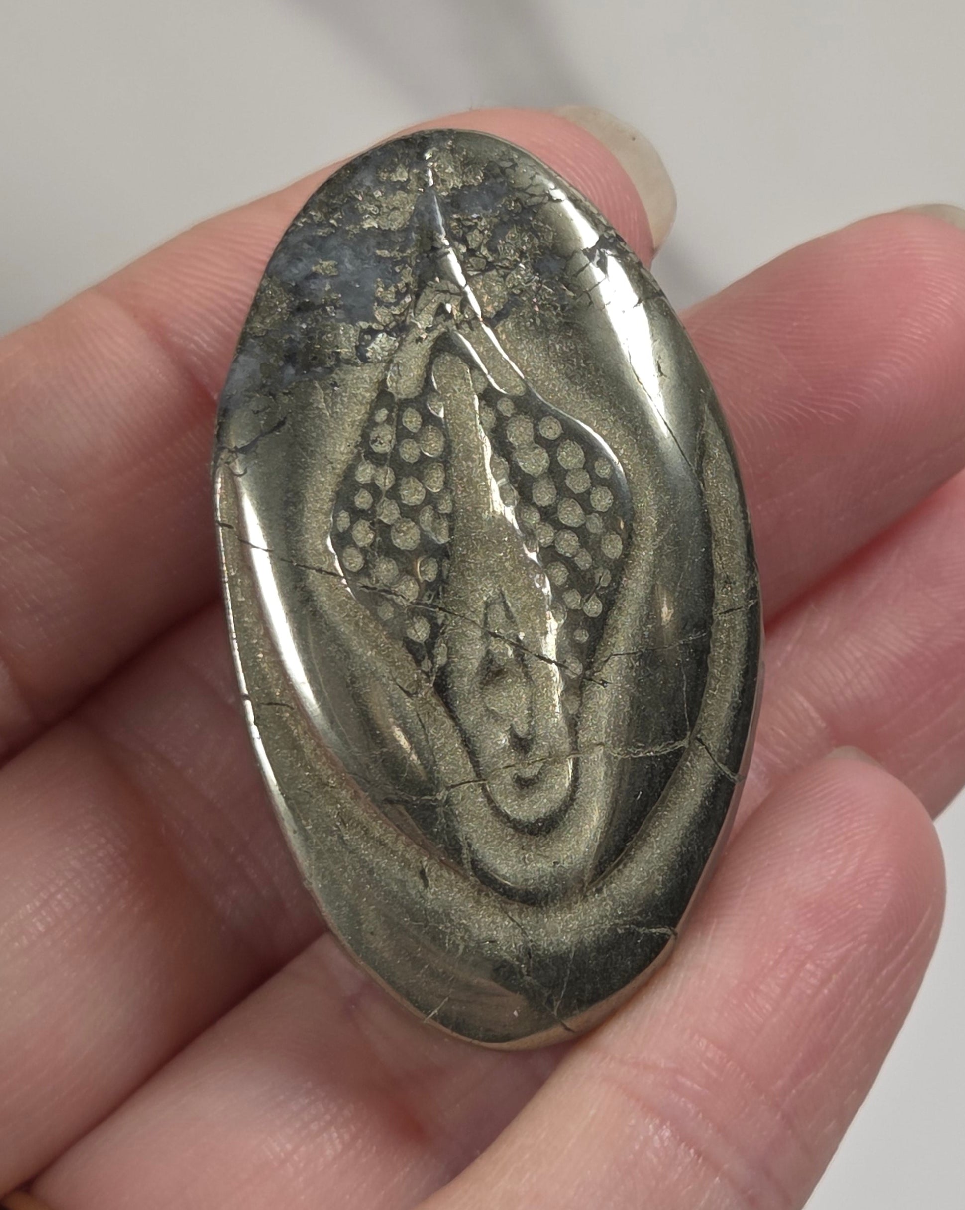 A hand holding an oval polished stone featuring a detailed fossil or reptile-like inlay, shown against a light marble-patterned background.