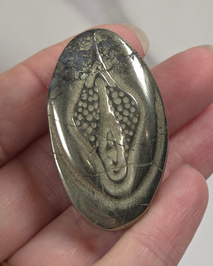 A hand holding an oval polished stone featuring a detailed fossil or reptile-like inlay, shown against a light marble-patterned background.
