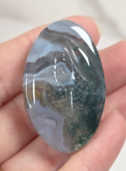 An oval polished gemstone with swirling gray, white, and dark green agate-like patterns, held in a person's hand.