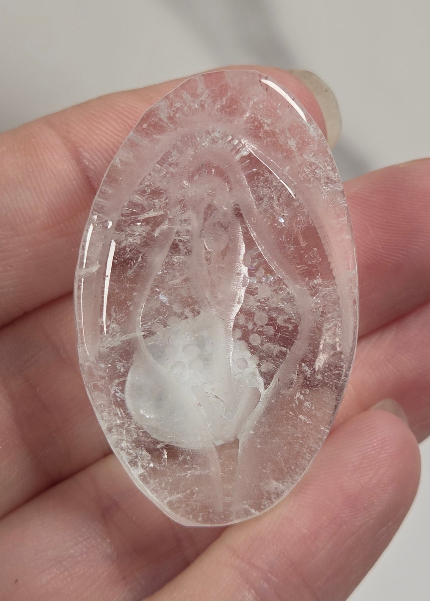 A hand holding a polished oval clear stone cabochon featuring a faint carved figure, with a gold ring on one finger against a soft light gray background.