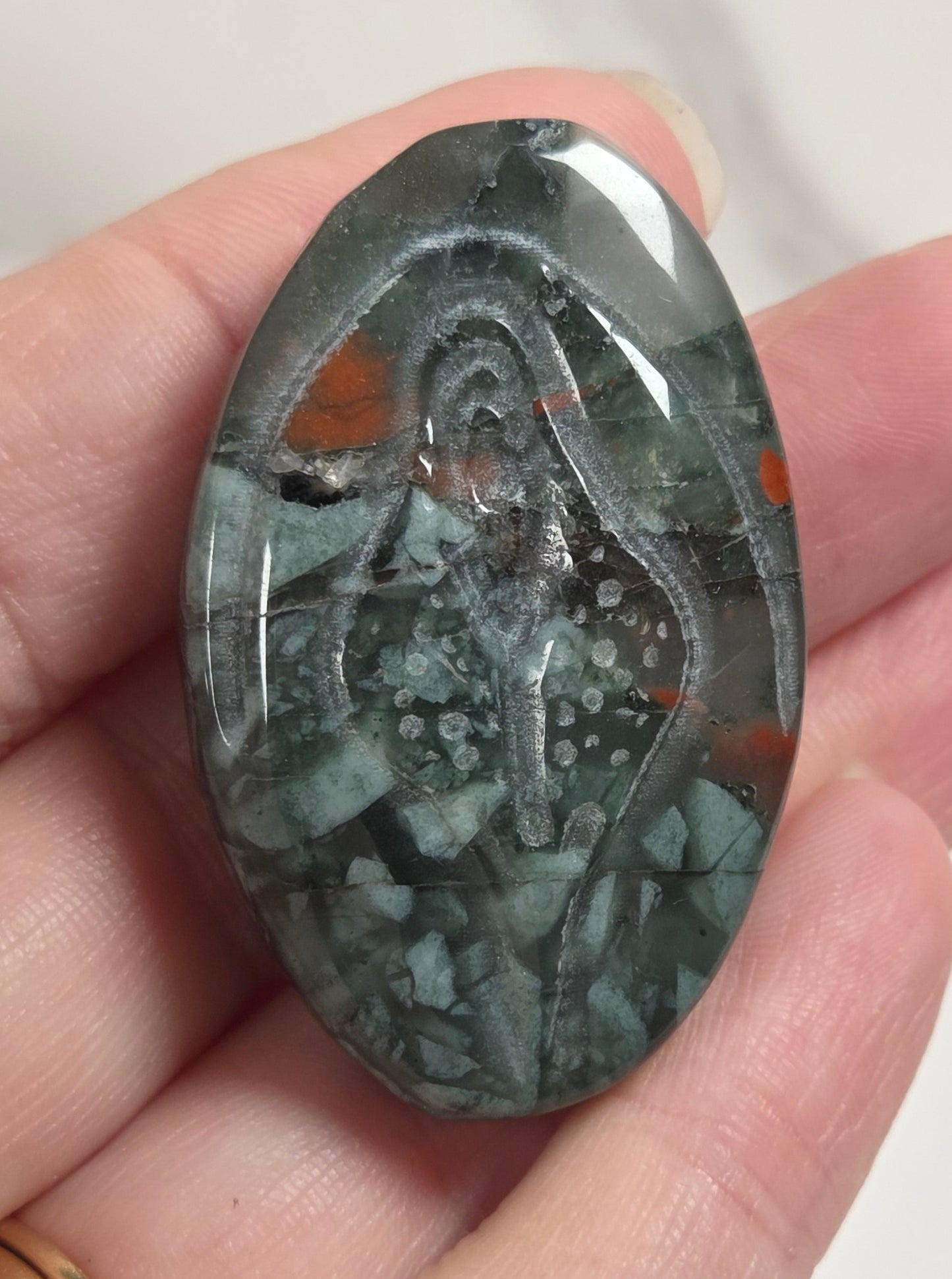A hand holds an oval polished gemstone cabochon featuring a carved figure design in dark gray, green, and orange marbled stone.