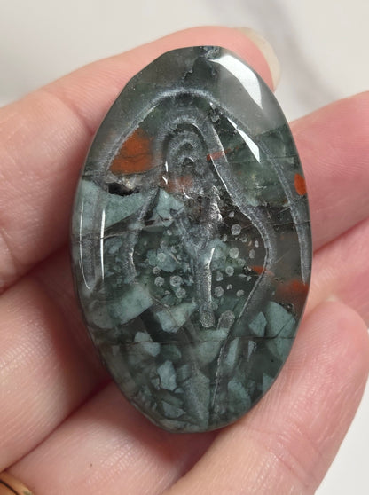 A hand holds an oval polished gemstone cabochon featuring a carved figure design in dark gray, green, and orange marbled stone.