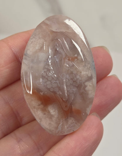 A hand holding a polished oval agate stone cabochon with natural dendritic patterns in soft pink, beige, and light brown tones.