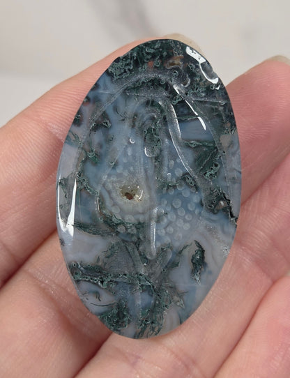 A hand holding an oval polished gemstone cabochon featuring a mottled pattern of dark green, gray, and white.