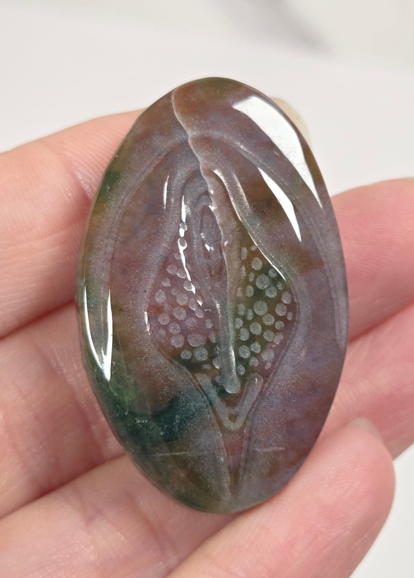 A hand holds a polished, oval-shaped carved stone cabochon featuring a stylized lotus flower design with green, purple, and brown marbled tones.