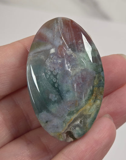 A hand holding a polished oval gemstone cabochon with a marbled pattern of teal, purple, gray, and gold hues, set against a light gray marble background.