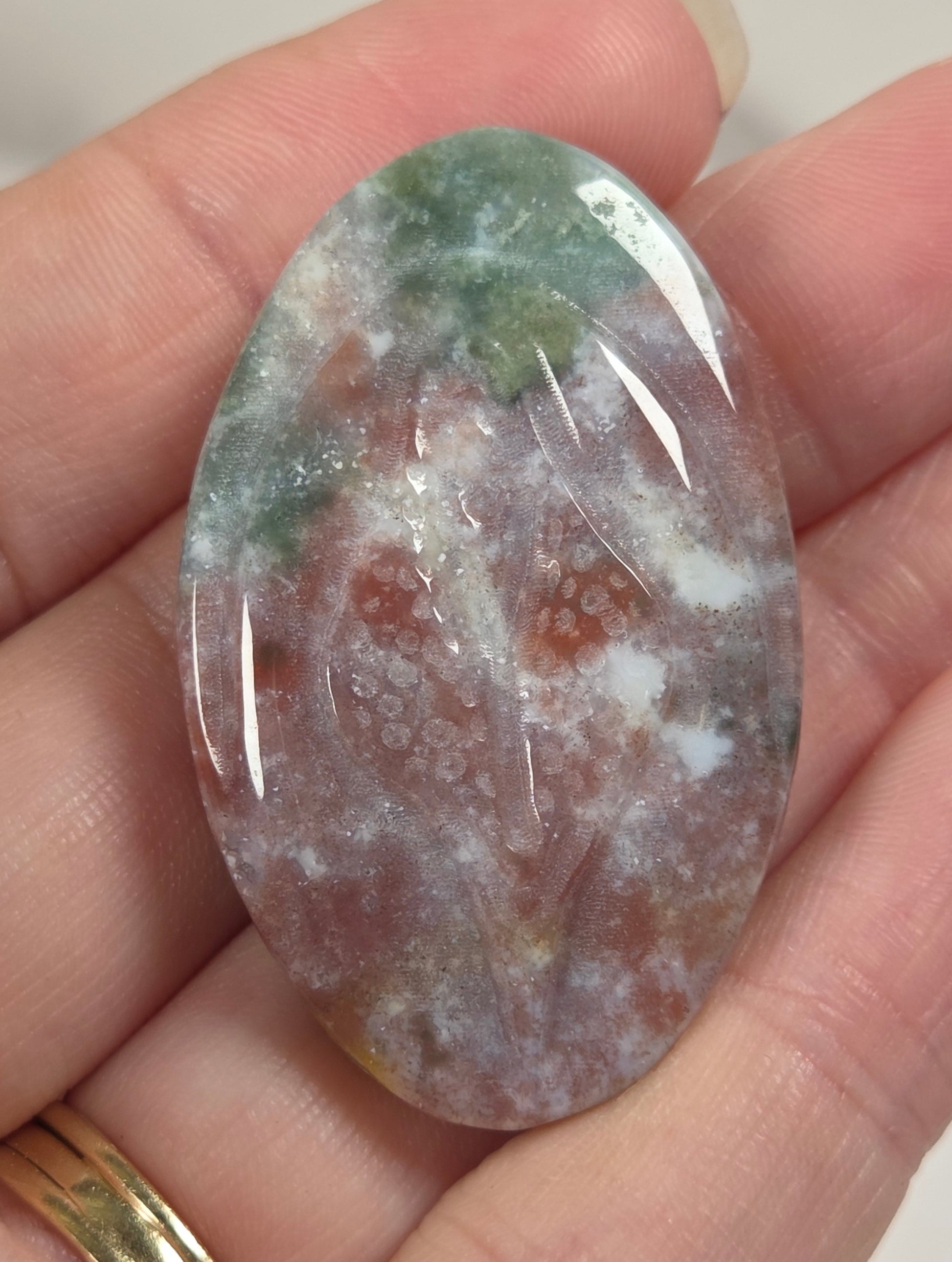 A hand holding a polished oval gemstone cabochon with a marbled pattern of green, white, and reddish-brown tones.