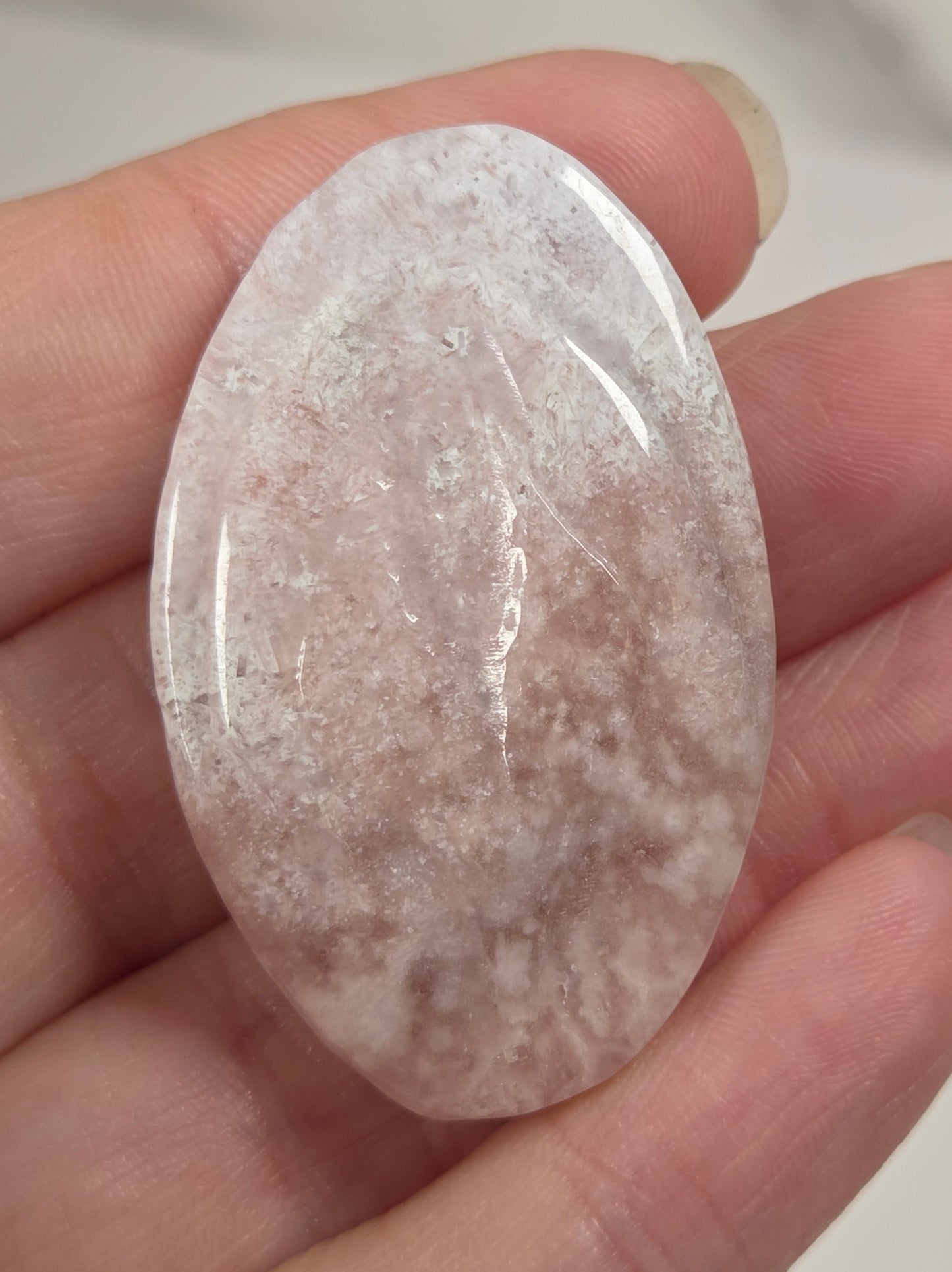 A hand holding a polished oval-shaped rose quartz crystal cabochon with natural inclusions and cloudy white and pinkish tones.