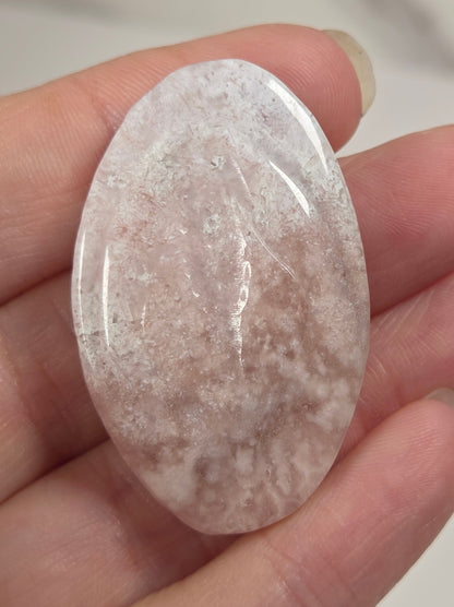 A hand holding a polished oval-shaped rose quartz crystal cabochon with natural inclusions and cloudy white and pinkish tones.