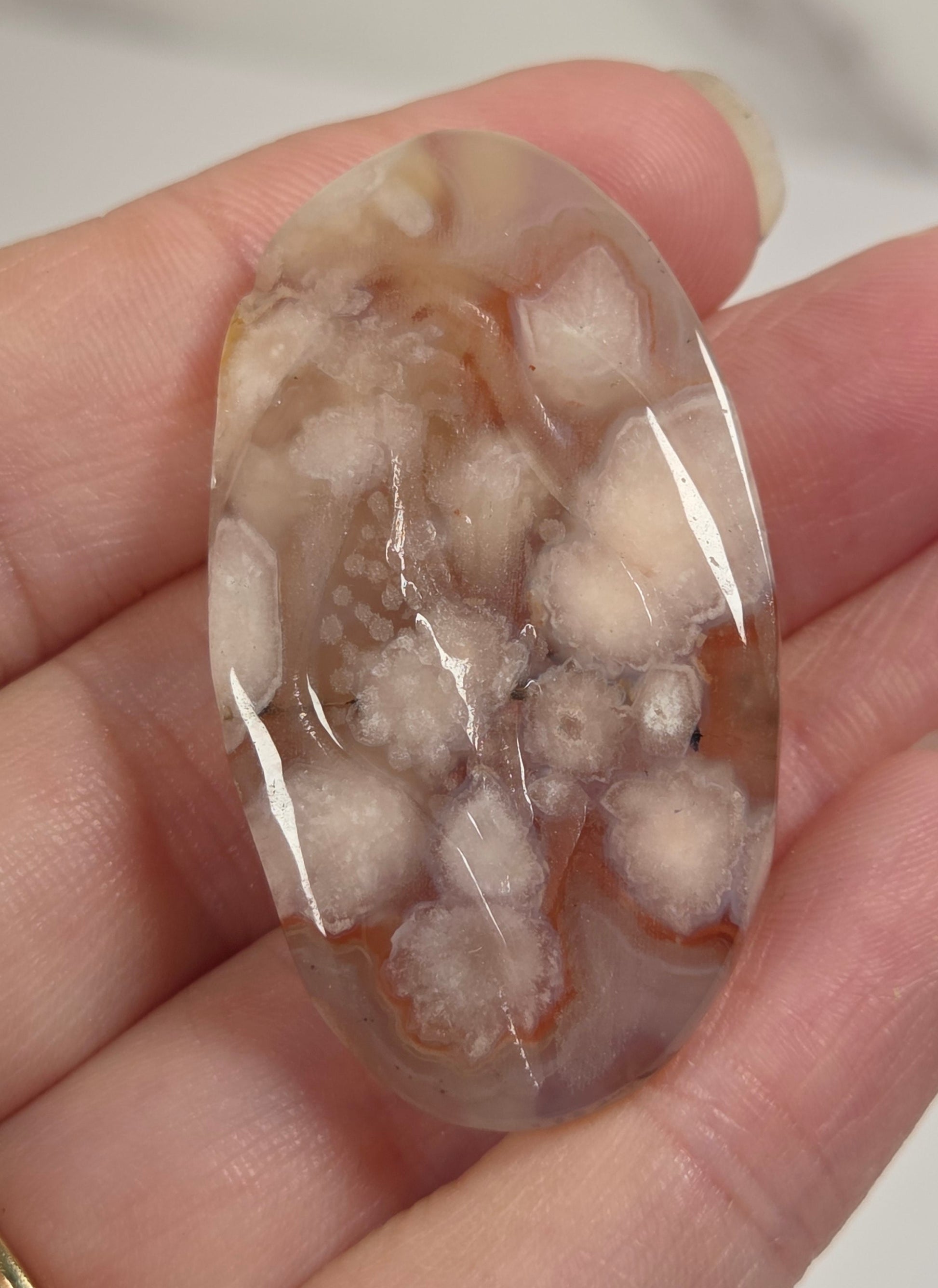A hand holding a polished oval cabochon gemstone with a marbled pattern of white, beige, and light brown, with a gold band ring visible on the finger.