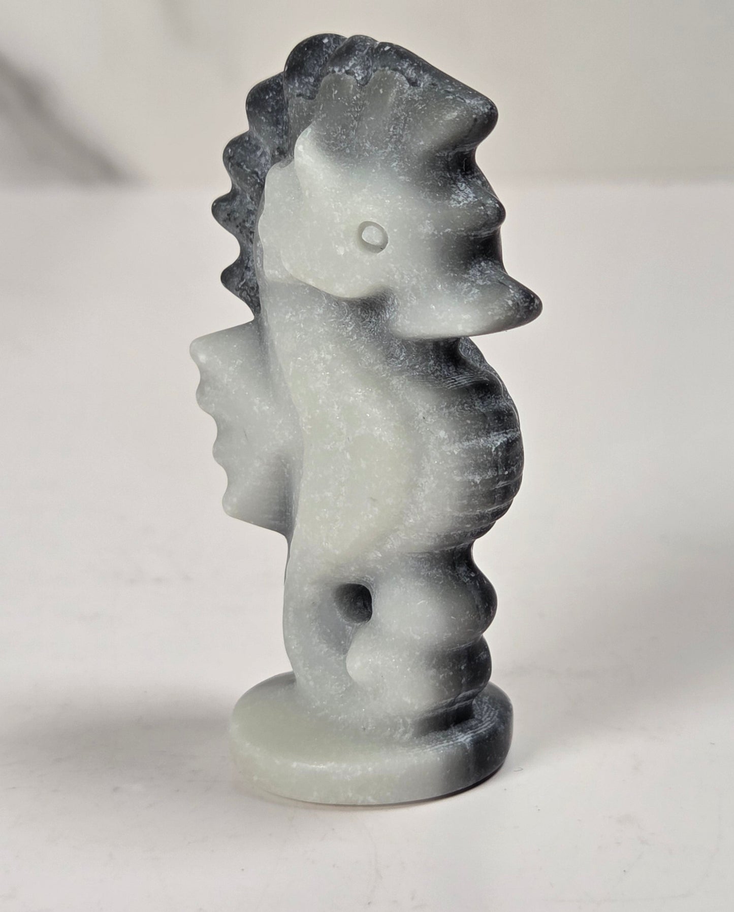A small carved seahorse figurine made of white stone with dark gray to black marbling, standing on a round base against a light gray background.