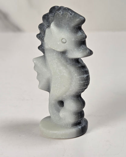 A small carved seahorse figurine made of white stone with dark gray to black marbling, standing on a round base against a light gray background.