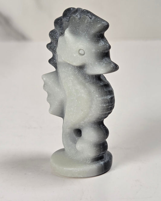 A small carved seahorse figurine made of white stone with dark gray to black marbling, standing on a round base against a light gray background.