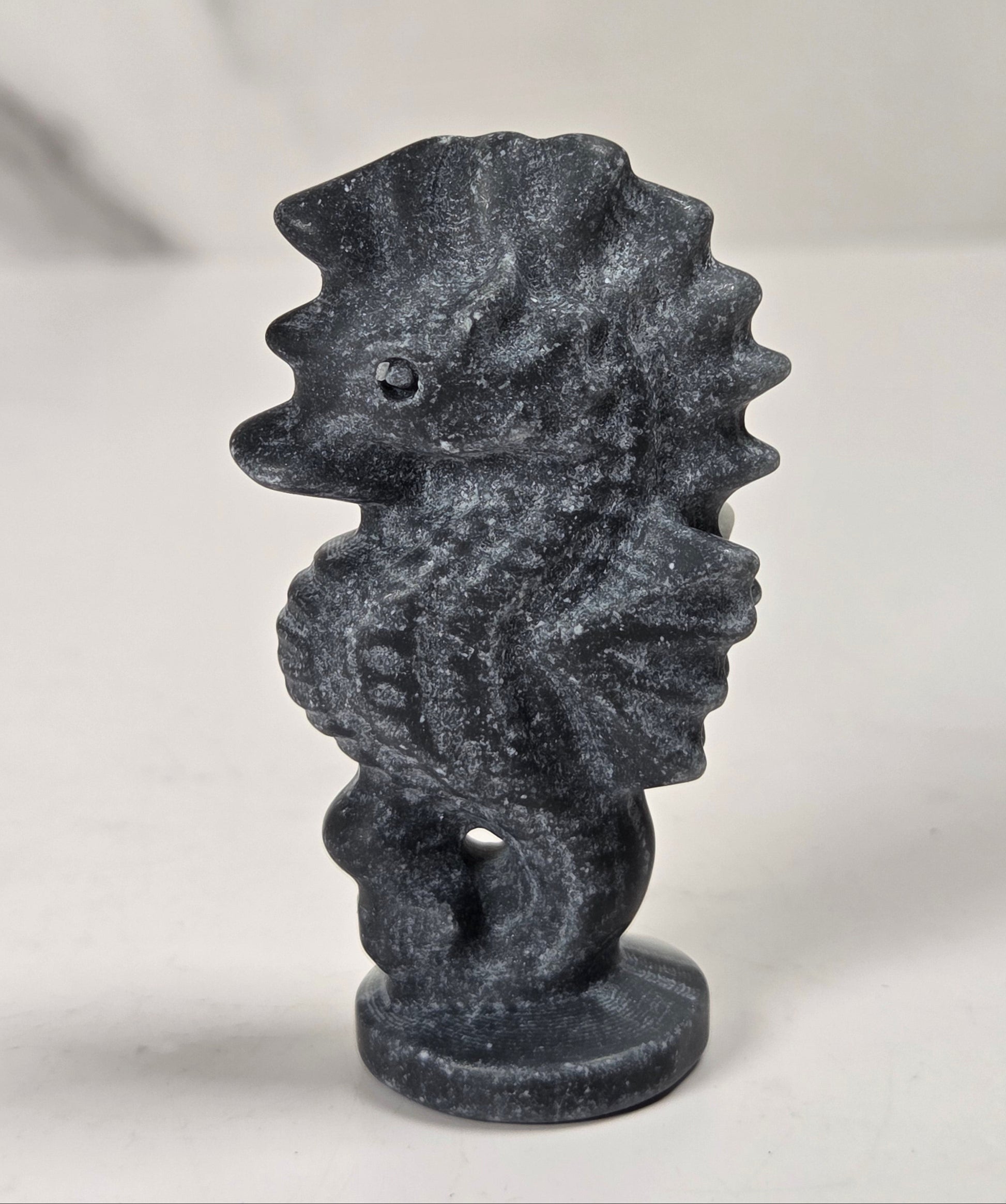 A small dark gray seahorse figurine with a textured, stone-like finish, standing on a round base against a light gray background.